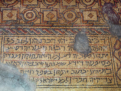 Ancient mosaic floor featuring intricate geometric patterns and a Hebrew inscription, with some sections damaged or worn.