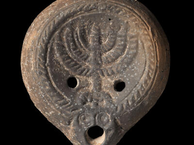 Ancient oil lamp with a decorative border and a central menorah design, with two holes flanking the menorah and a larger hole at the spout.