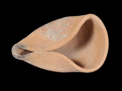 A clay oil lamp with a wide, rounded body and a pointed spout, showing surface wear and some discoloration.