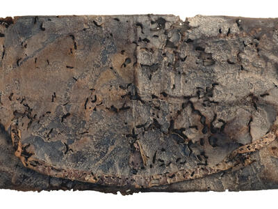 A folded rectangular piece of weathered, decayed leather with holes, dark stains, and a frayed strap or cord along one edge.