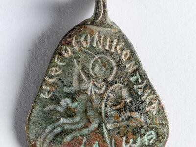 A weathered metal object with a triangular shape, featuring faint engraved text and an image of a horse and rider. The surface shows signs of age and patina, with a hook or loop at the top.