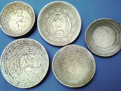Five ancient ceramic bowls with inscriptions in a Semitic script, written in concentric circles. Some bowls feature geometric shapes and human-like figures.