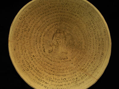 A shallow, round clay bowl with an inscription in a Semitic script written in concentric circles surrounding a central drawing of a human-like figure.