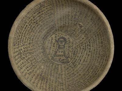 A round, ancient clay bowl inscribed with lines of text, some concentric and some radial, in a dark Semitic script. In the center is a simple drawing of a figure with wide eyes, surrounded by additional writing.