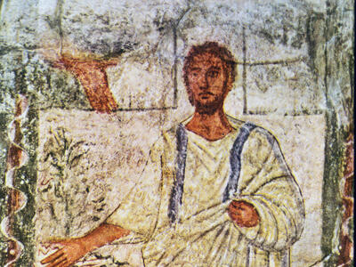 Ancient fresco shows a bearded man in a white robe with blue stripes, standing beside a tree, extending one arm outward. The background features simple landscape shapes, and a wavy border surrounds the scene.