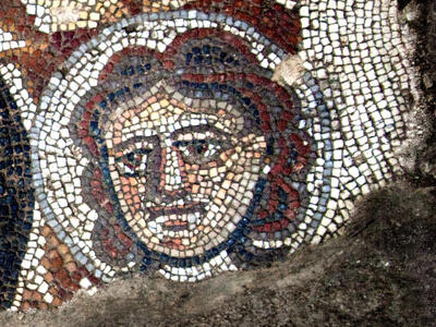 Ancient mosaic depicting a human face with red hair, large eyes, and a neutral expression, made from small colored tiles. The face is outlined with a halo-like border.