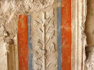 A partially damaged ancient wall features red and blue vertical stripes with a raised relief of a leafy vine in the center, set against a textured, weathered surface.