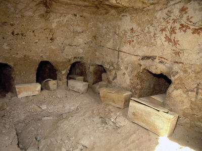 Ancient stone ossuaries rest on a dusty cave floor, with multiple burial niches carved into the wall and faint frescoes of grape vines visible on the rough stone surface.
