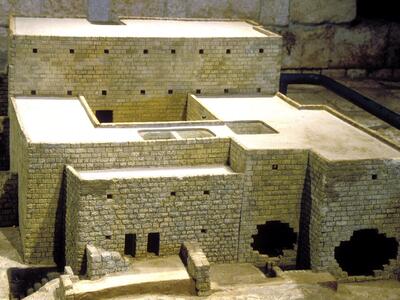 A detailed scale model of an ancient stone building featuring rectangular walls and small window openings, displayed against a stone backdrop.