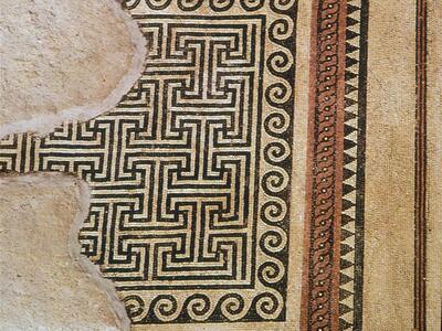 Ancient mosaic floor with geometric patterns, including a black-and-white labyrinth design, surrounded by colored borders; part of the mosaic is damaged and missing on the left side.