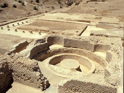 Ancient ruins with stone walls and a circular structure, set in a desert landscape with barren hills in the background under a clear blue sky.