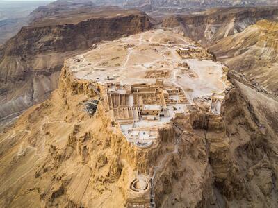 Aerial view of an ancient fortress, perched atop a rugged, rocky plateau in a desert landscape with scattered ruins and surrounding mountains.