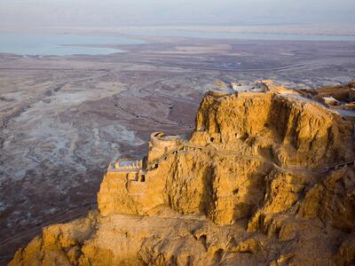 Aerial view of an ancient fortress atop a rugged desert plateau, with dramatic cliffs leading down to the dry landscape and the Dead Sea visible in the background.