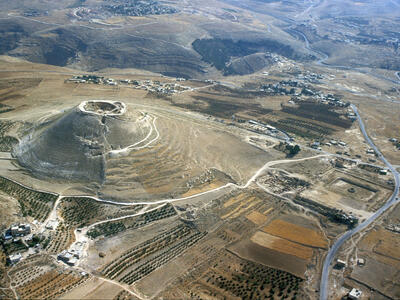Aerial view of a rounded, steep-sided hill with ruins on top, surrounded by arid land, fields, scattered trees, roads, and small settlements in a hilly landscape.