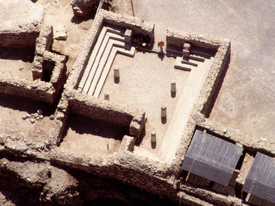 Aerial view of an ancient stone structure with steps along the sides, five central pillars, and partially intact surrounding walls in a desert archaeological site.