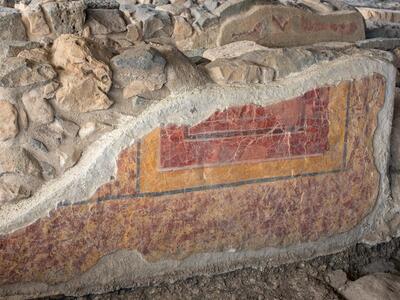 Ancient stone wall with a preserved red, orange, and yellow geometric fresco, surrounded by rough stones and partially eroded plaster.