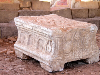 A carved stone object with detailed reliefs, including menorahs and arches, resting on four short legs, sits on a dirt floor among weathered stone blocks and ruins.