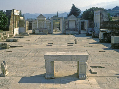 Ancient stone ruins of a large hall with mosaic floors and carved columns, open to the sky, surrounded by trees and hills in the background. At the near end of the hall is a two-legged table flanked by the remains of animal statues. At the far end is a doorway between two niches with triangular tops.