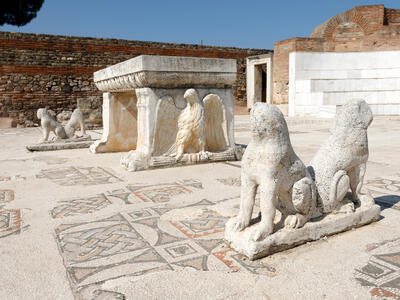 Ancient marble statues of lions surround a rectangular table with a relief of an eagle visible on its side. The table and statues rest on a mosaic-tiled floor, set in a partially preserved brick and stone courtyard under a clear sky. Behind them is a semicircular structure with stadium-like benches.