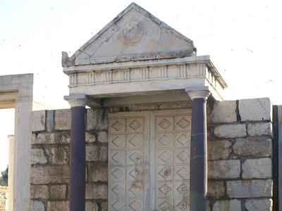 A stone structure with steps leading to a closed double door, flanked by columns and topped with a triangular pediment, set against a wall of large rectangular stones.