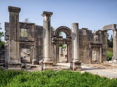 Partially preserved stone building facade with an arched entrance and four columns in front. Other stone ruins are visible through the entryway.