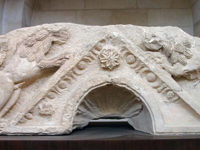 A weathered stone fragment featuring two carved lions surrounding a triangular roof with an arched opening.