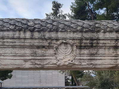 A stone lintel rests atop two stone pillars, set outdoors with trees and a modern building visible in the background. In the center of the lintel is a relief of a menorah surrounded by a wreath, with faint Hebrew inscriptions to its right and left.
