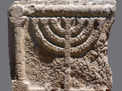 Ancient stone relief depicting a seven-branched menorah with a base, carved in raised detail, flanked by a column on the left.