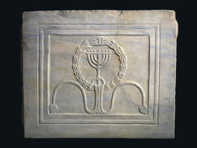 A carved marble tablet featuring a menorah inside a wreath, with two curved lines ending in leaf shapes beneath it. The design is framed by a rectangular border.