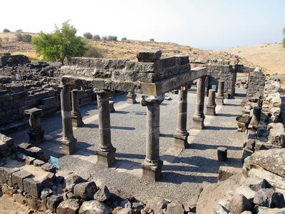 Ancient stone ruins of a rectangular structure with several columns and partial walls, set in a dry, hilly landscape with sparse vegetation under a clear sky.