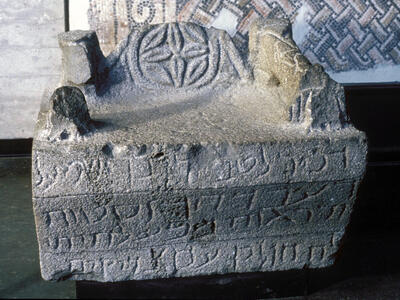 Ancient stone chair with carved Hebrew inscriptions and a circular symbol on the backrest, displayed in a museum setting with mosaic artwork visible in the background.