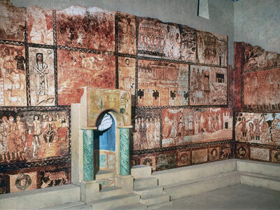Colorful ancient wall paintings with biblical scenes and figures surround a small, elaborately decorated niche with stairs, set against a stone floor and walls in an archaeological restoration.