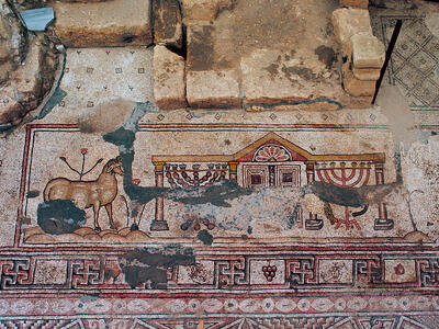 Ancient mosaic with a deer, a building with columns, menorahs, ritual objects, and geometric patterns, partially damaged and surrounded by stone ruins.