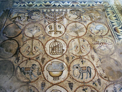 Ancient mosaic featuring interlocking circles with animals, trees, a caged bird, grapes, and geometric patterns, bordered by a zigzag design and two lions facing each other, flanking a menorah, at the top.