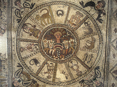Ancient mosaic featuring a central figure surrounded by twelve sections, each depicting a zodiac sign with symbolic animals or figures, bordered by decorative patterns and human faces.