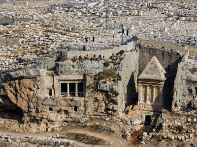 An ancient rock-cut tomb complex with rectangular openings and a pyramid-roofed structure stands in a vast hillside cemetery with countless stone graves and a few people.