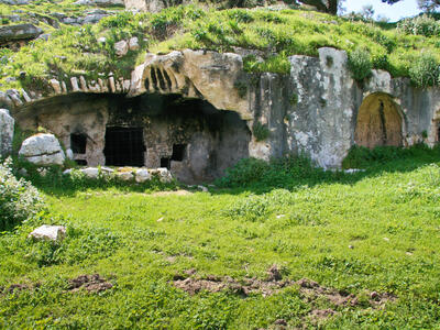Ancient rock-cut tombs set into a grassy hillside, with arched entrances and stone walls partially covered by green vegetation. One entrance is barred with metal grates.