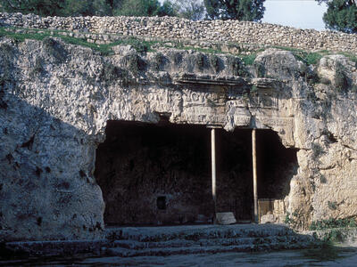 A rock-cut tomb with ancient carvings above the entrance, two stone columns, and steps leading up. A stone wall and trees are visible on the hill above the tomb.