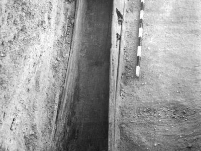 Black-and-white photo of a wooden coffin in an excavated grave site, seen from above, with a measuring stick placed beside it for scale and a sandbag at one end.