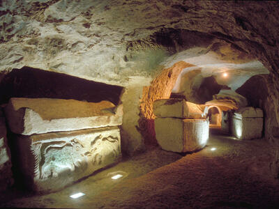 Ancient underground stone passage with carved sarcophagi, illuminated by soft lights; textured rock walls and ceiling create a cave-like atmosphere.