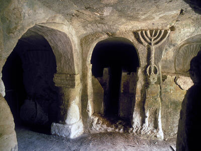 Ancient stone chamber with arched doorways and carved reliefs, including a prominent menorah, on the walls. The space is dimly lit, showing textured surfaces and worn details.