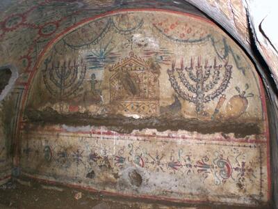 Ancient wall painting in a stone tomb featuring ornate menorahs, a depiction of a Torah shrine or ark, decorative patterns, and symbolic objects. The artwork is faded and partially damaged.