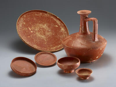 A collection of ancient red-glazed pottery, including a large jug with a tall neck and handle, a wide shallow dish, and four smaller dishes.
