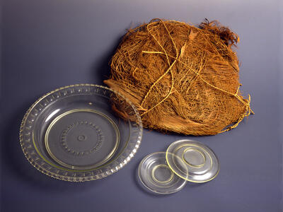 A bundle of palm fiber mat, a round clear glass dish, and two small clear glass dishes are arranged on a smooth, dark surface.