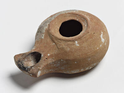 A round, brown ancient clay oil lamp with a spout for a wick, showing black burn marks and white residue.