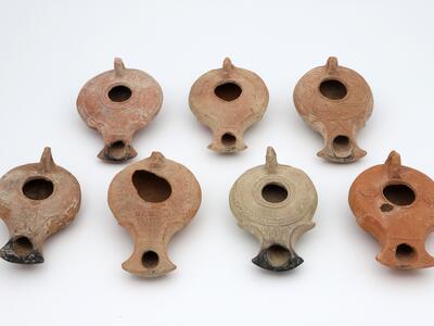 Seven ancient clay oil lamps are arranged on a white surface. Each lamp has a round body, a central hole, a small handle, and a spout, with varying shades of brown and beige and some visible decorative patterns.
