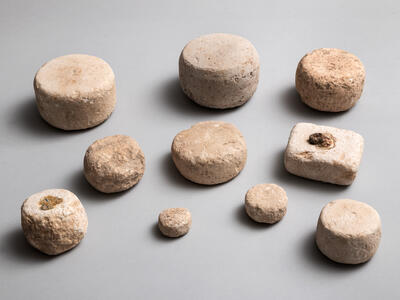 Ten ancient stone weights of various sizes and shapes, mostly cylindrical and one roughly rectangular, arranged on a plain gray background. The stones appear worn and textured, with earthy beige tones.