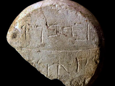 A broken, round stone with ancient script carved into its rough surface, featuring two visible lines of inscriptions, against a black background.