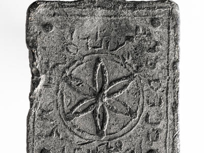 A rectangular, weathered metal object with a central flower-like geometric pattern inside a circle, surrounded by worn Hebrew inscriptions, with a hole at the top for hanging.