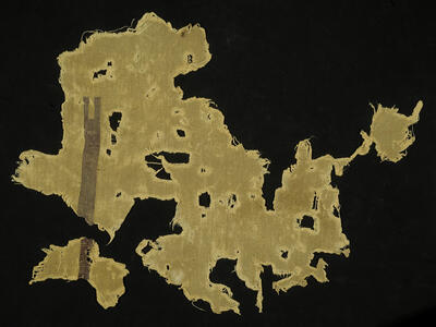 A yellowish, irregularly shaped ancient textile fragment with frayed edges and multiple holes, featuring a verticle stripe with a notch at the end.